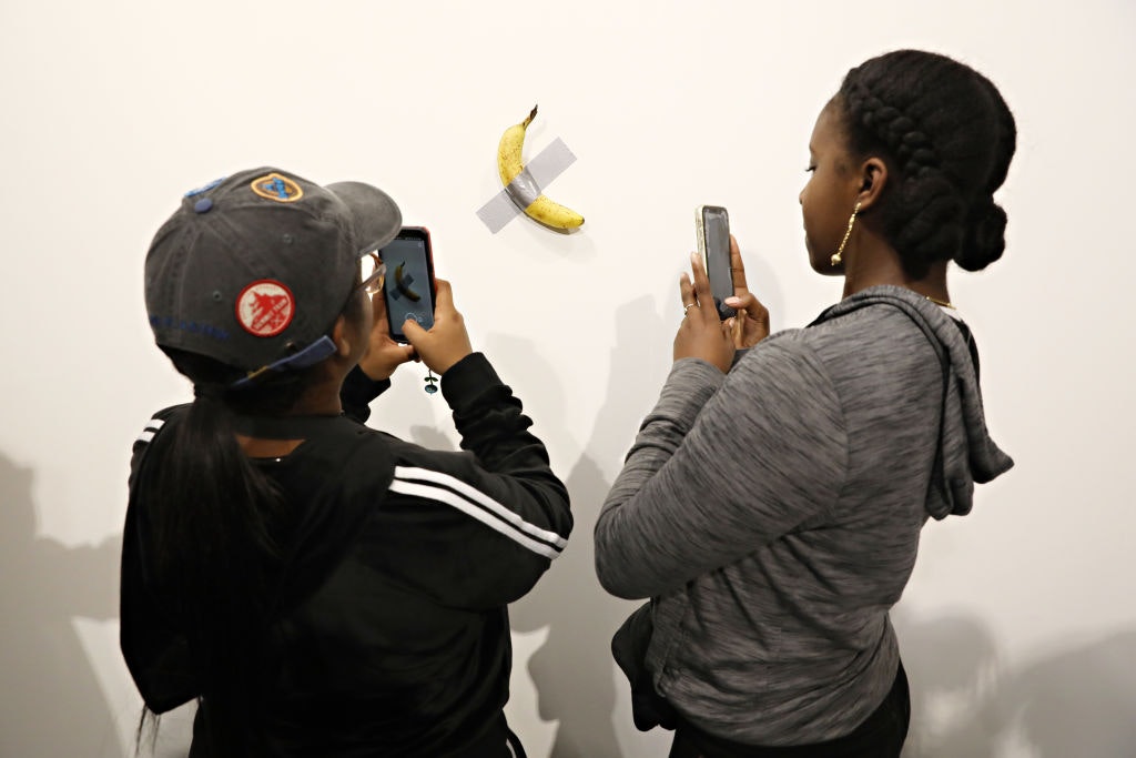 İki kişi akıllı telefonlarıyla duvara bantlanmış bir muzun fotoğrafını çekiyor.